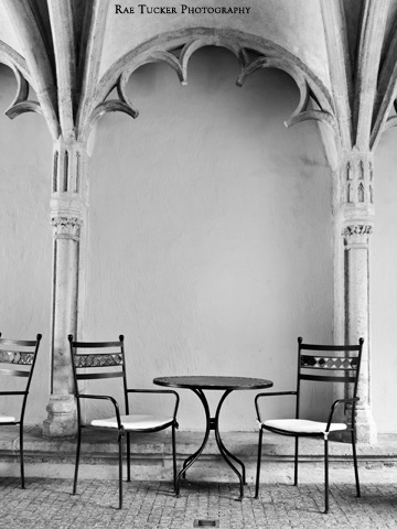 A black and white image of a patio table and chairs under a stone arch.