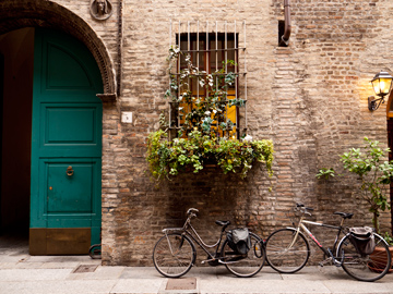 A brick wall in Parma, Italy hosts a turquoise door, bicycles, plants and a window