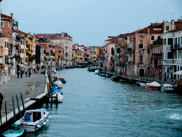 A larger canal is lined with boats and pedestrians in Venice, Italy